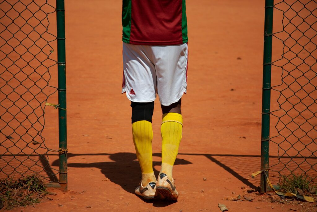 Rear view of a soccer player wearing a red and green soccer jersey, white shorts, yellow socks, and cleats on a clay field, demonstrating how to style a soccer jersey for on-field performance. Image by RODOLFO BARRETTO via www.unsplash.com